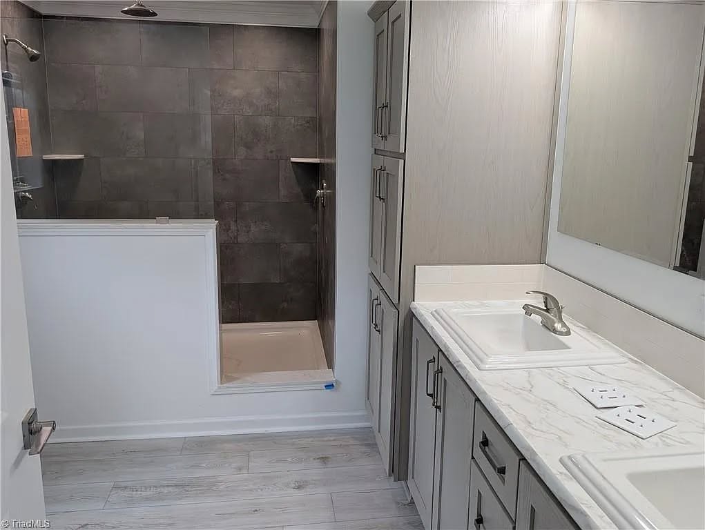 Modern bathroom with gray tiled shower, half wall, dual sinks on a white marble countertop, light wood cabinets, and a large mirror. Clean and minimalist.
