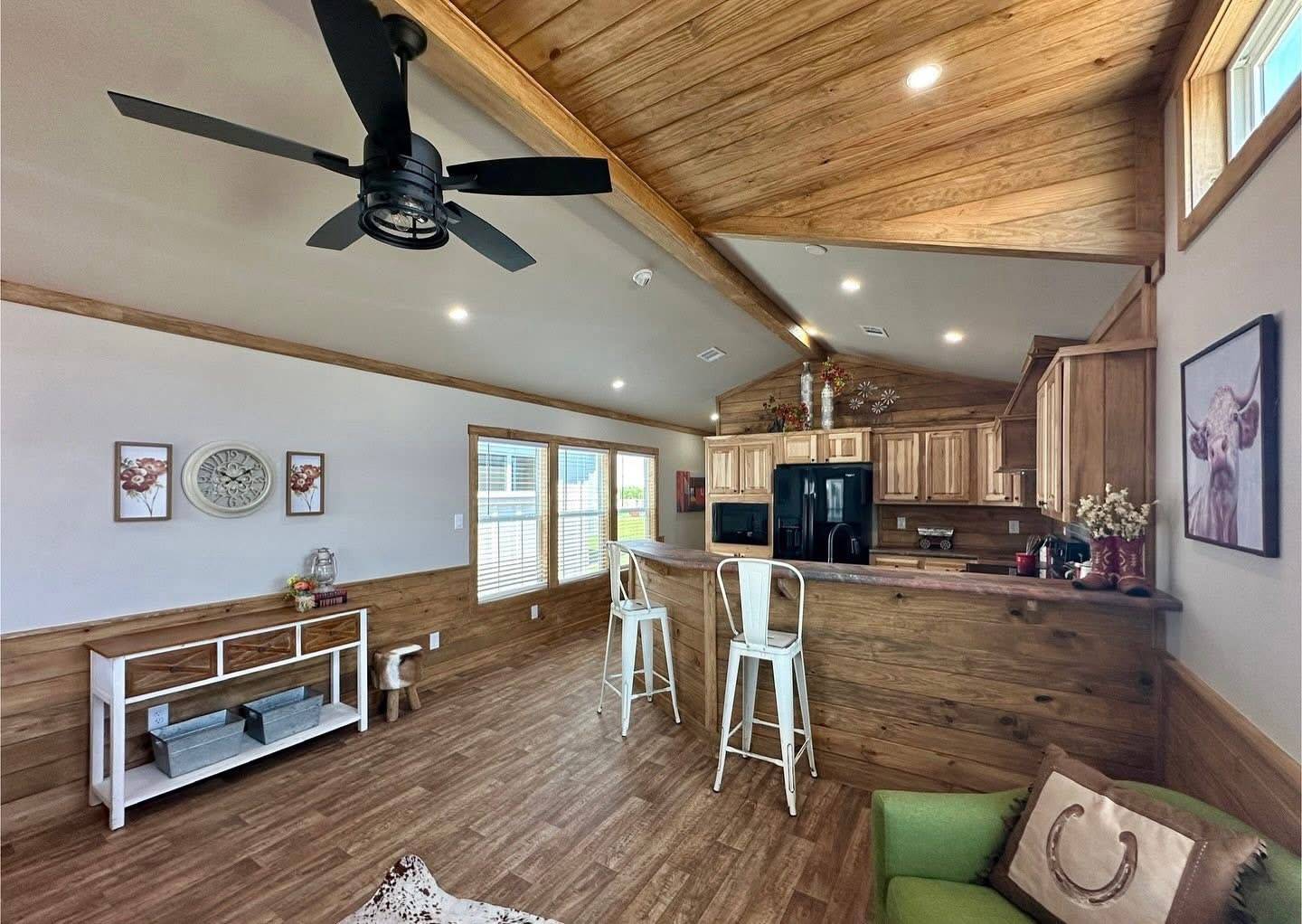 A cozy living area with wood accents features a ceiling fan, bar stools at a wooden counter, and a wall clock. Bright natural light fills the space.