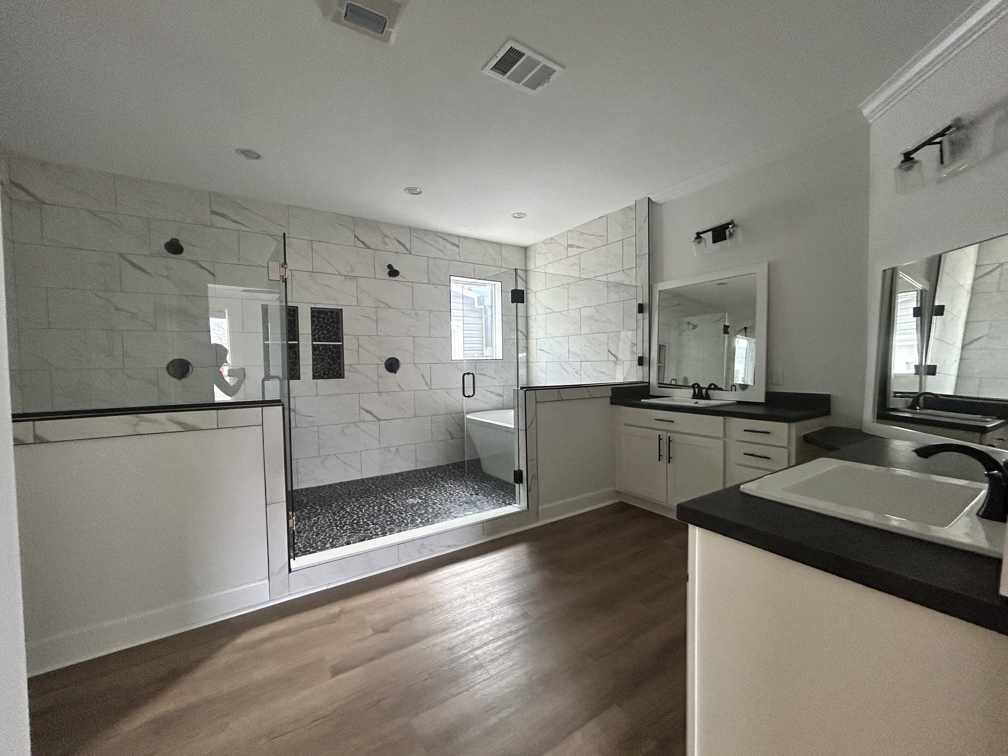 Modern bathroom with a large glass shower, white tiles, and black accents. Features a freestanding bathtub, large mirror, and wood-style flooring.
