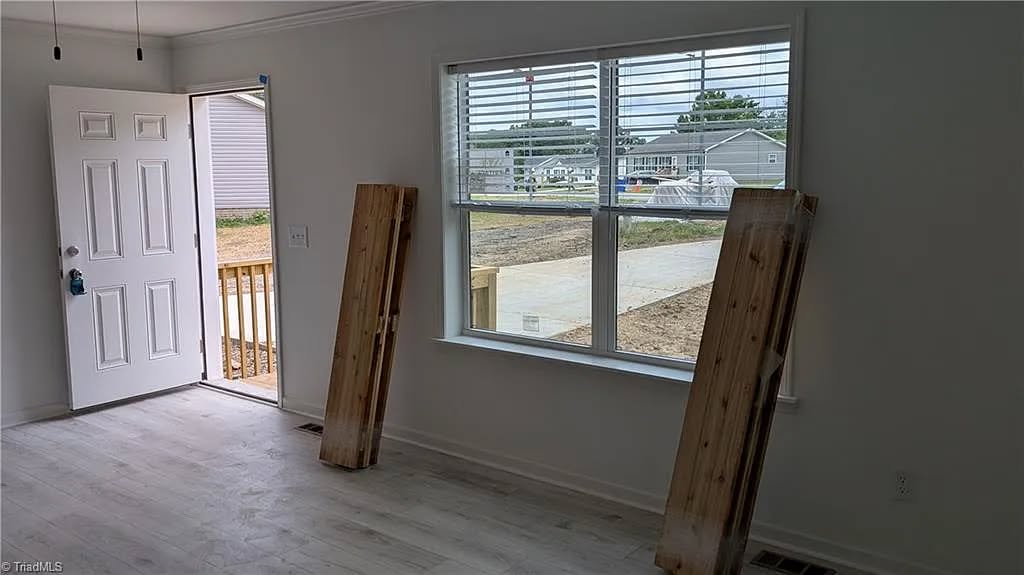 A bright, empty room with white walls and light wood flooring. An open door leads outside, with sunlight streaming in. Two wooden planks are propped by a window overlooking a construction area. The atmosphere is minimal and fresh.