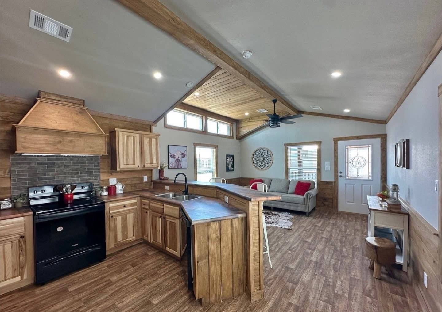 Rustic open-concept living room and kitchen with wood accents. Features a black stove, couch with red cushions, bar stools, and a ceiling fan. Cozy and inviting.