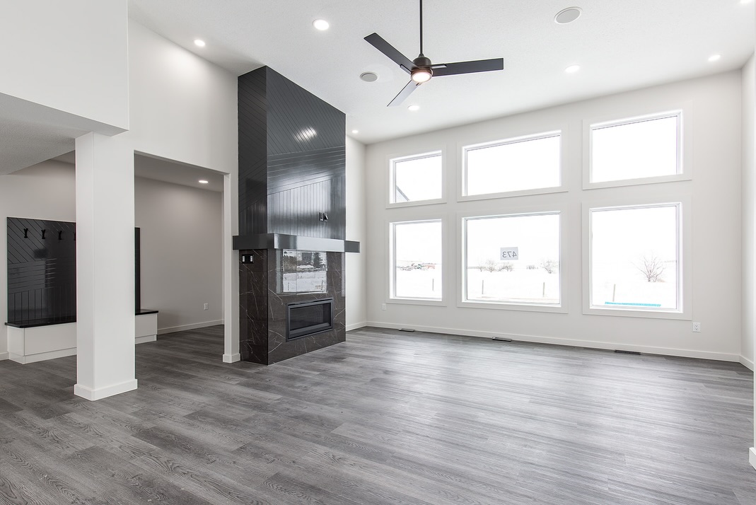 Spacious modern living room with large windows, gray wood flooring, black fireplace, high ceiling with fan, and abundant natural light.
