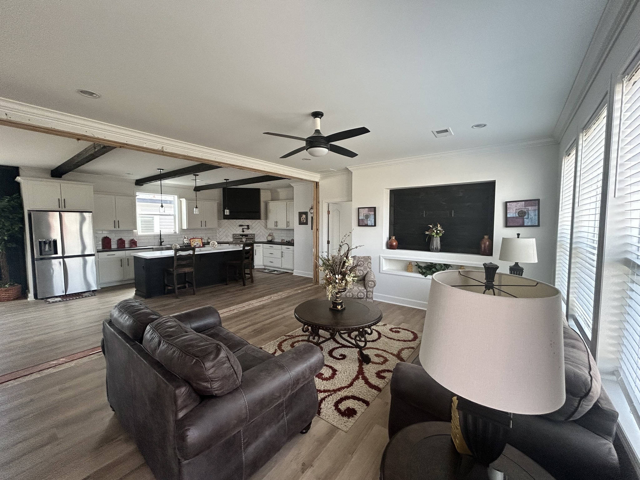 Spacious open-plan living area with gray couches, round coffee table, and stylish kitchen. Warm tones, decorative rug, and ample natural light.