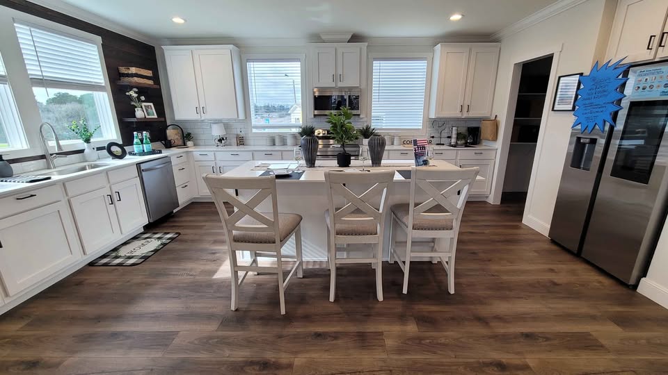 Modern kitchen with white cabinets and island, three beige cushioned chairs, stainless steel appliances, and dark wooden floor. Bright, cozy ambiance.