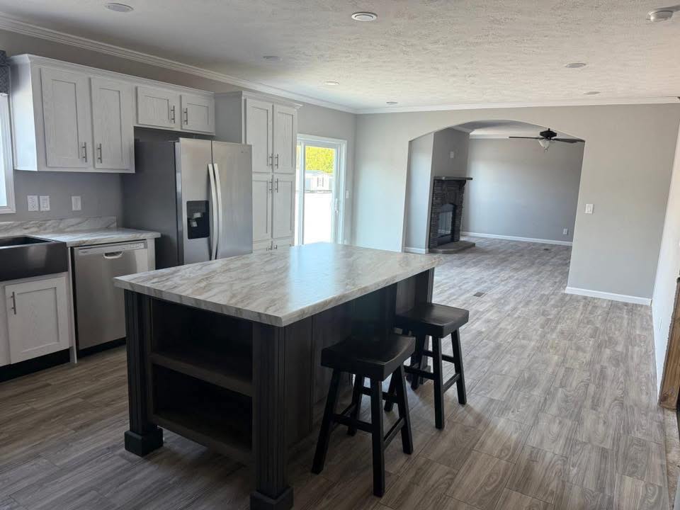 Modern kitchen with light gray cabinets, marble island, and stainless steel appliances. Wood flooring extends to a living area with a ceiling fan.