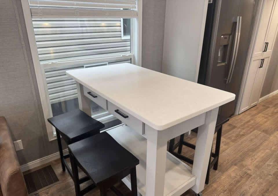 A modern kitchen with a white island table, four black stools, and a large window with blinds. A stainless steel refrigerator is nearby, set on wooden flooring.