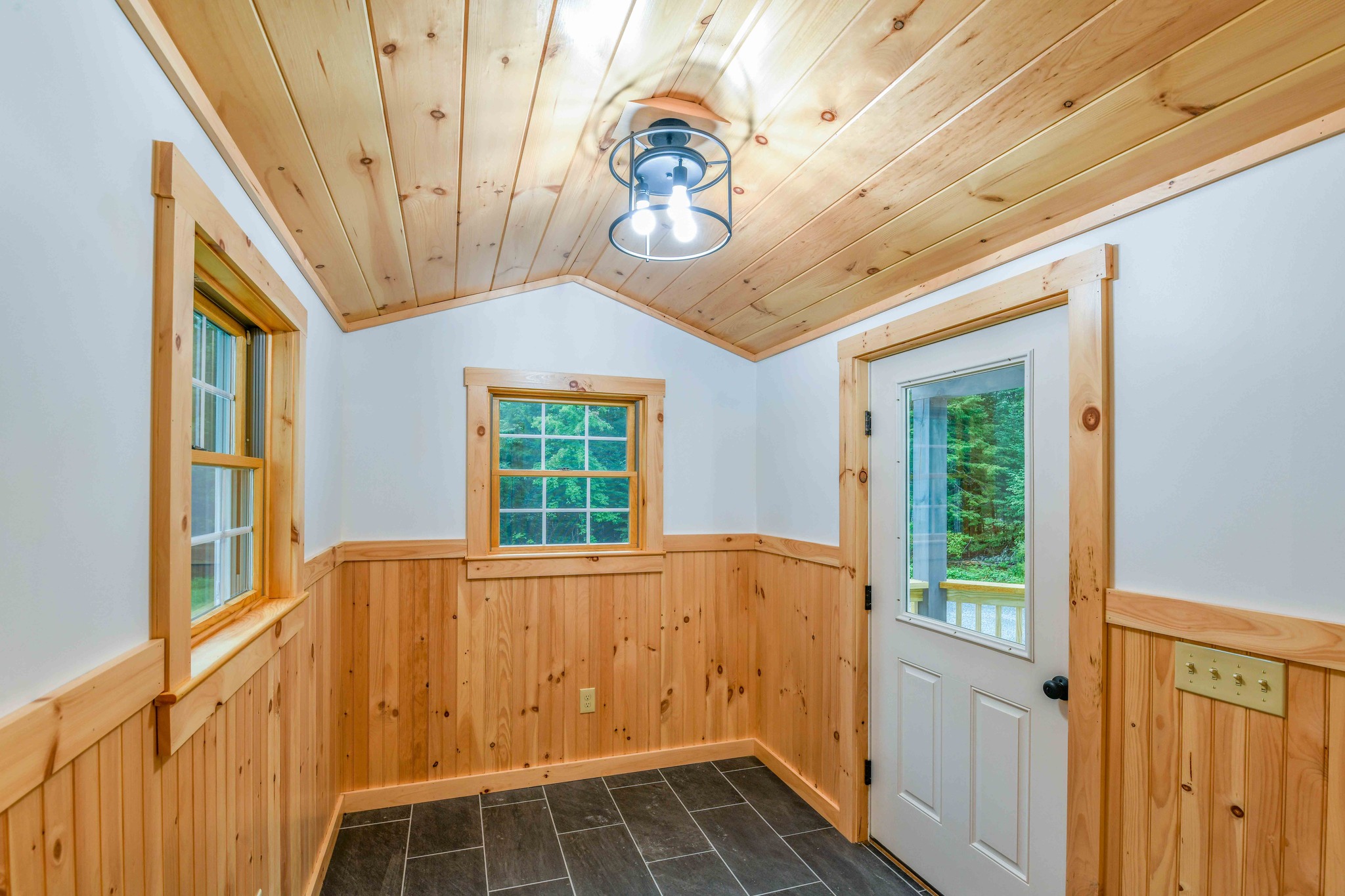 Wood-paneled entryway with dark slate tile floor, featuring a simple light fixture on the ceiling. A window and glass door overlook a green yard.
