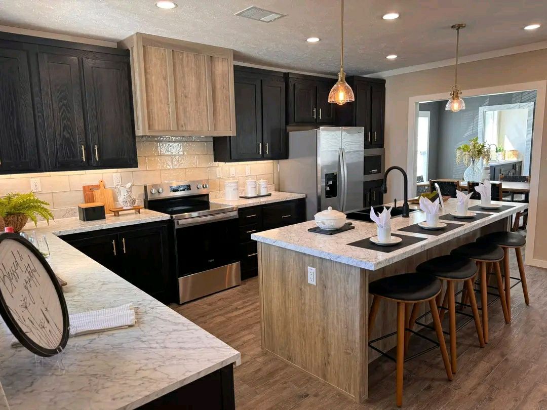 Modern kitchen with dark cabinets, light countertops, and stainless steel appliances. Central island with stools, stylish pendant lighting, and dining area visible.