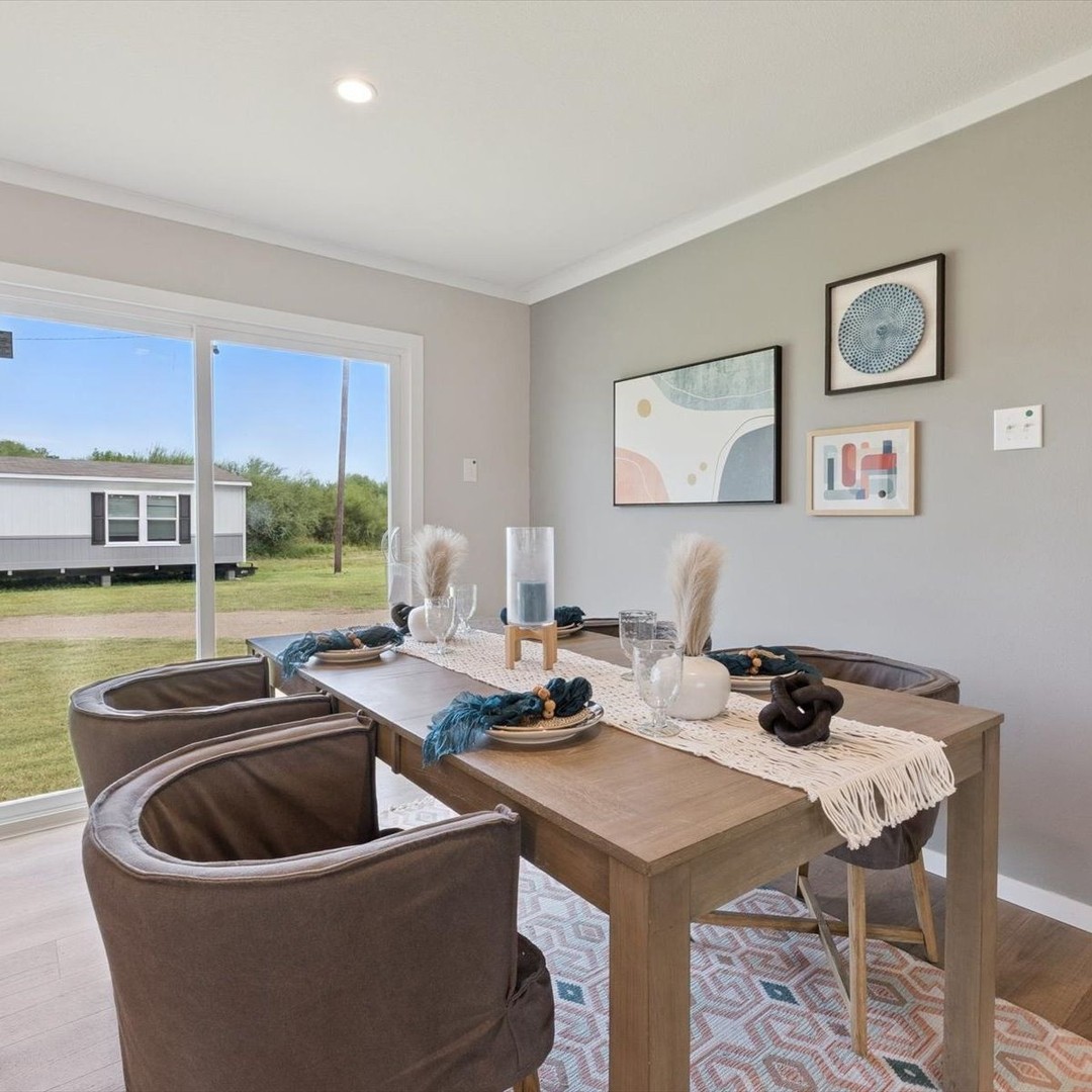 Modern dining room with a wooden table set for four, blue napkins, pampas grass centerpiece, leather chairs, abstract wall art, and a large window showing a green yard.