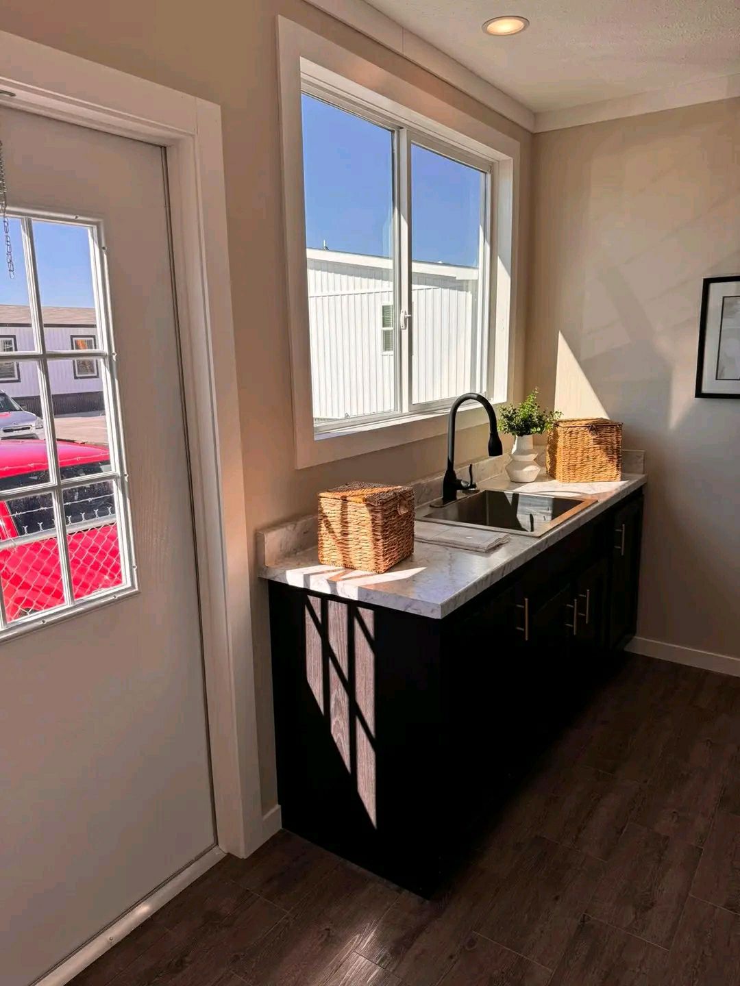 Compact kitchen space with a large window, white counter, black cabinets, and wicker baskets. Sunlight casts soft shadows. Calm and minimalist ambiance.