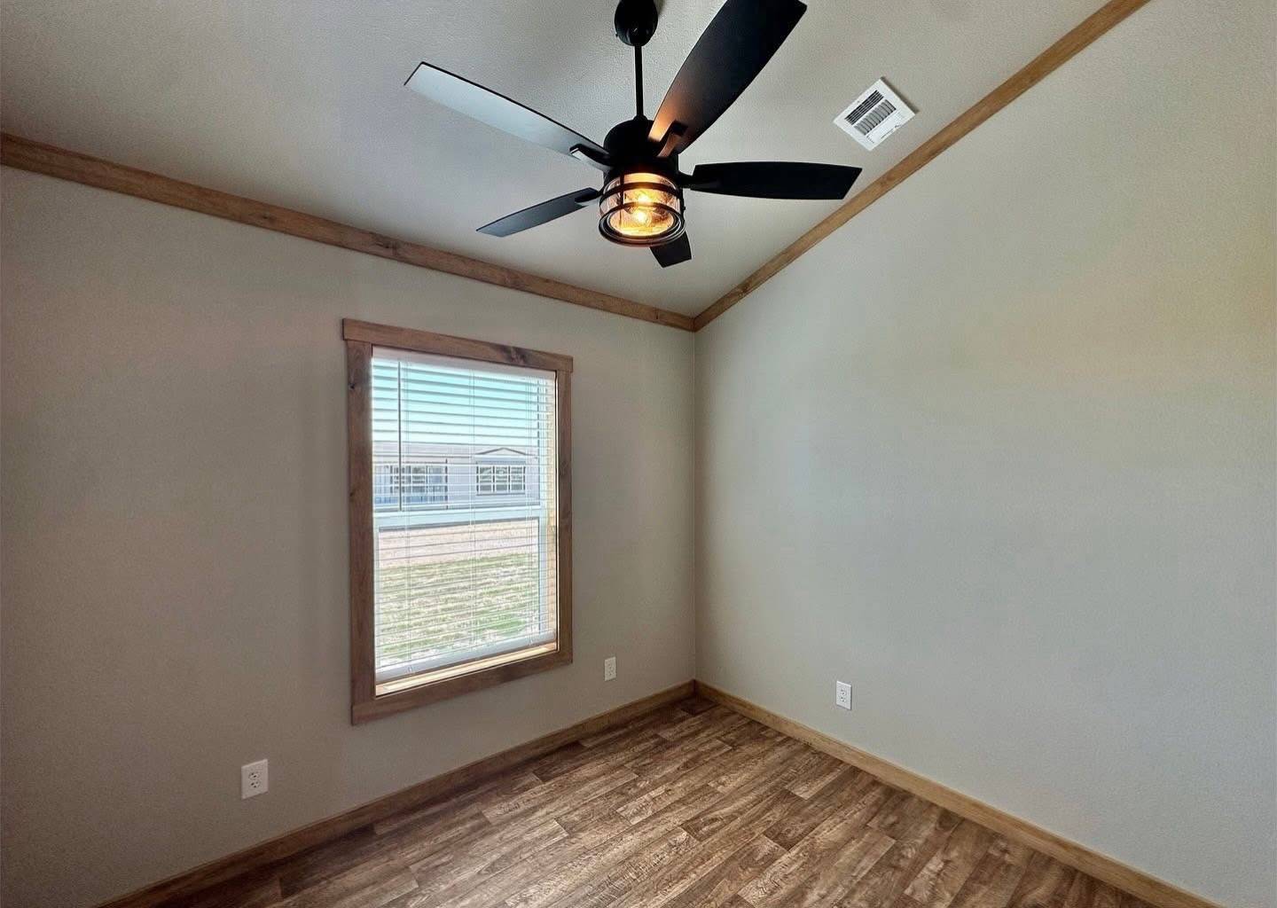 A small, empty room with beige walls and wooden trim, featuring a dark ceiling fan and a single window with blinds. The floor is wood-style.