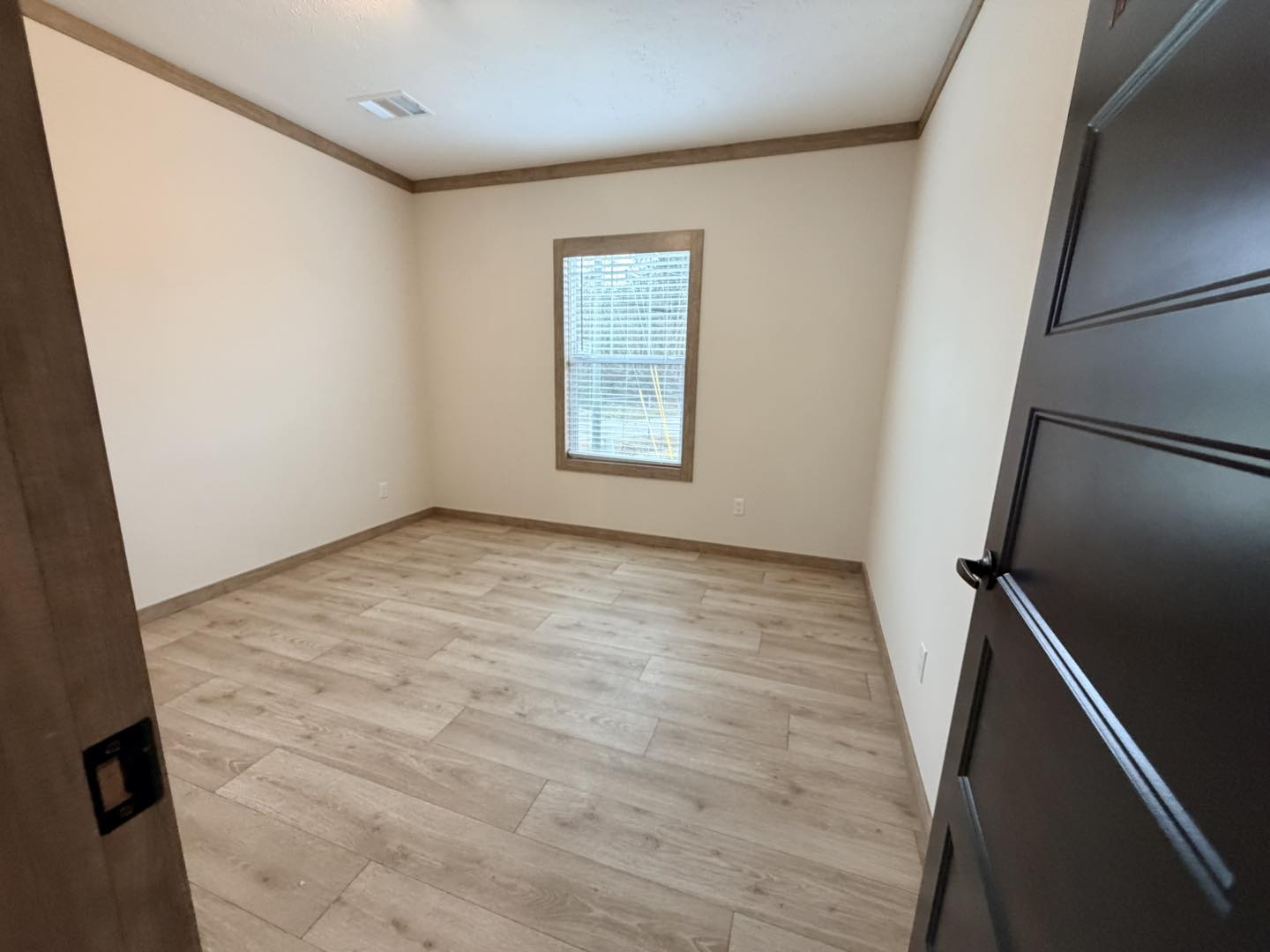 Empty room with light wood flooring, beige walls, and a single window with blinds. A dark wooden door is partially open, creating a minimalistic and inviting space.