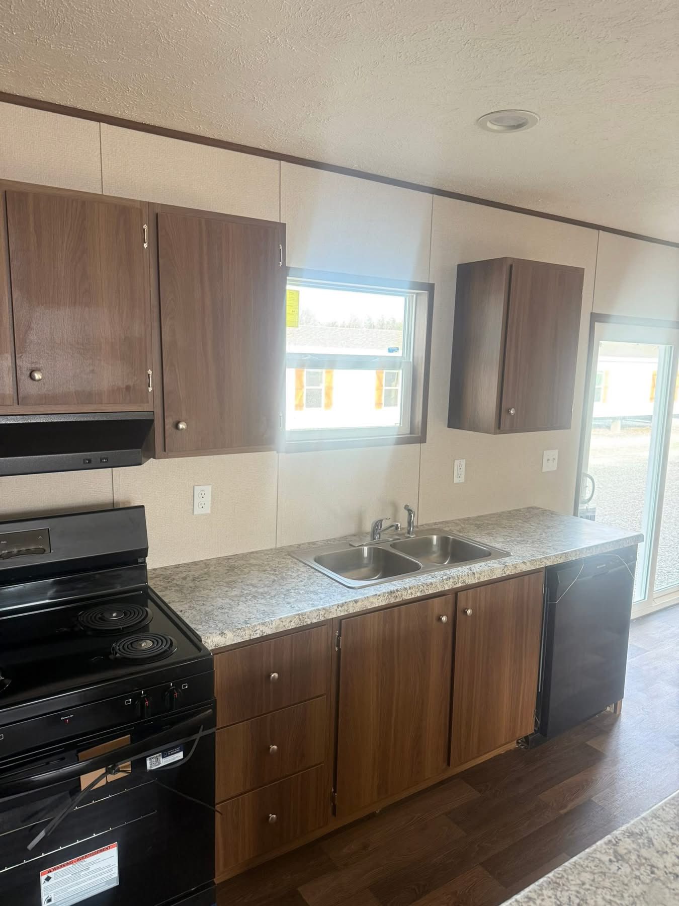 Compact kitchen with wooden cabinets, black stove, dual sinks, and granite countertops. Sunlight enters through a window, creating a warm, inviting atmosphere.