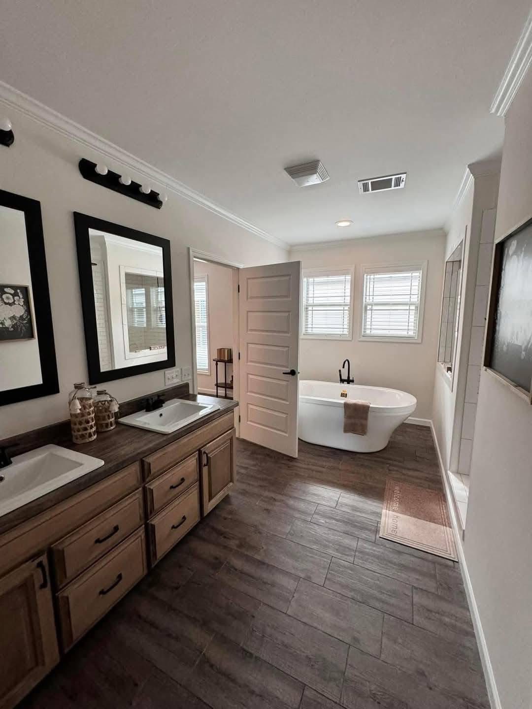 Spacious bathroom with wooden flooring, double sinks on a wooden vanity, two mirrors, and a freestanding bathtub by a window. Bright and serene atmosphere.