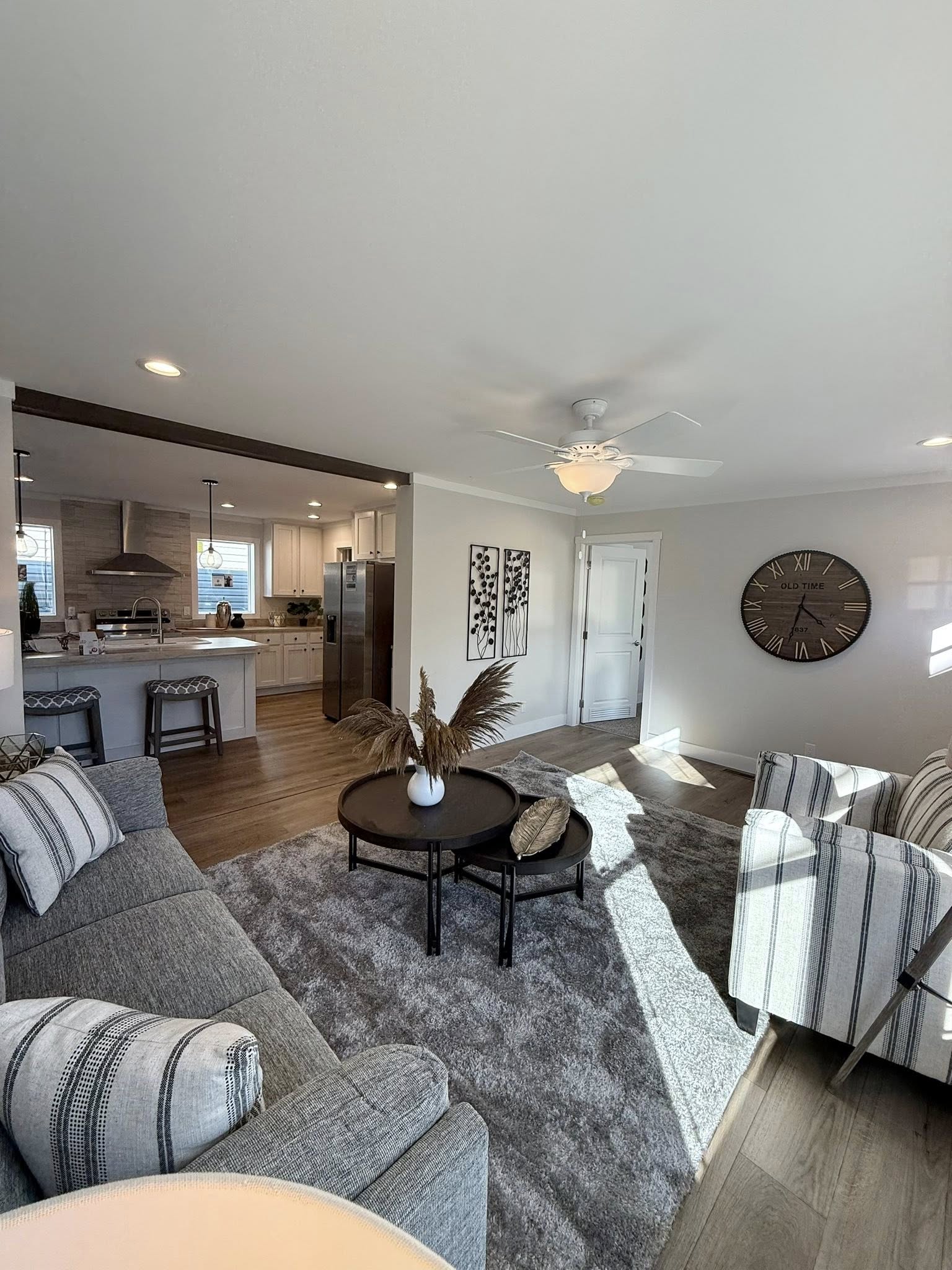 Modern living room with gray sofa, striped armchairs, and round coffee table on a plush rug. Wall clock and kitchen in background, bright and inviting.