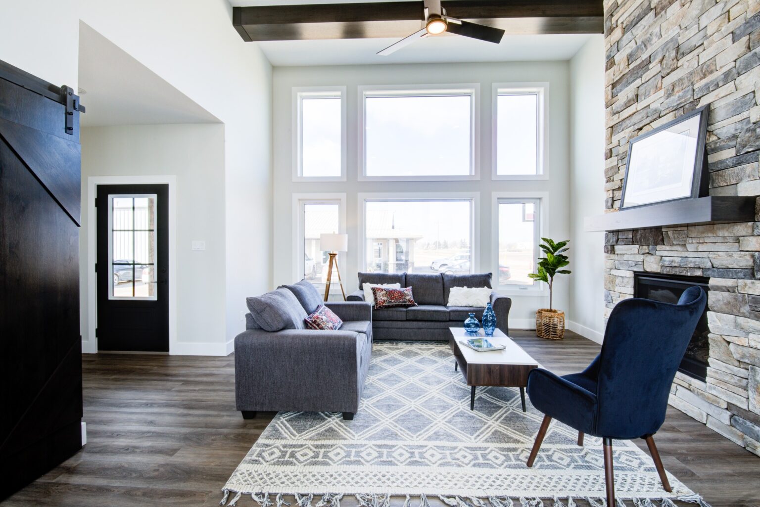 Bright, modern living room with large windows, stone fireplace, and dark wood accents. Cozy gray sofas, patterned rug, and a navy chair convey warmth.