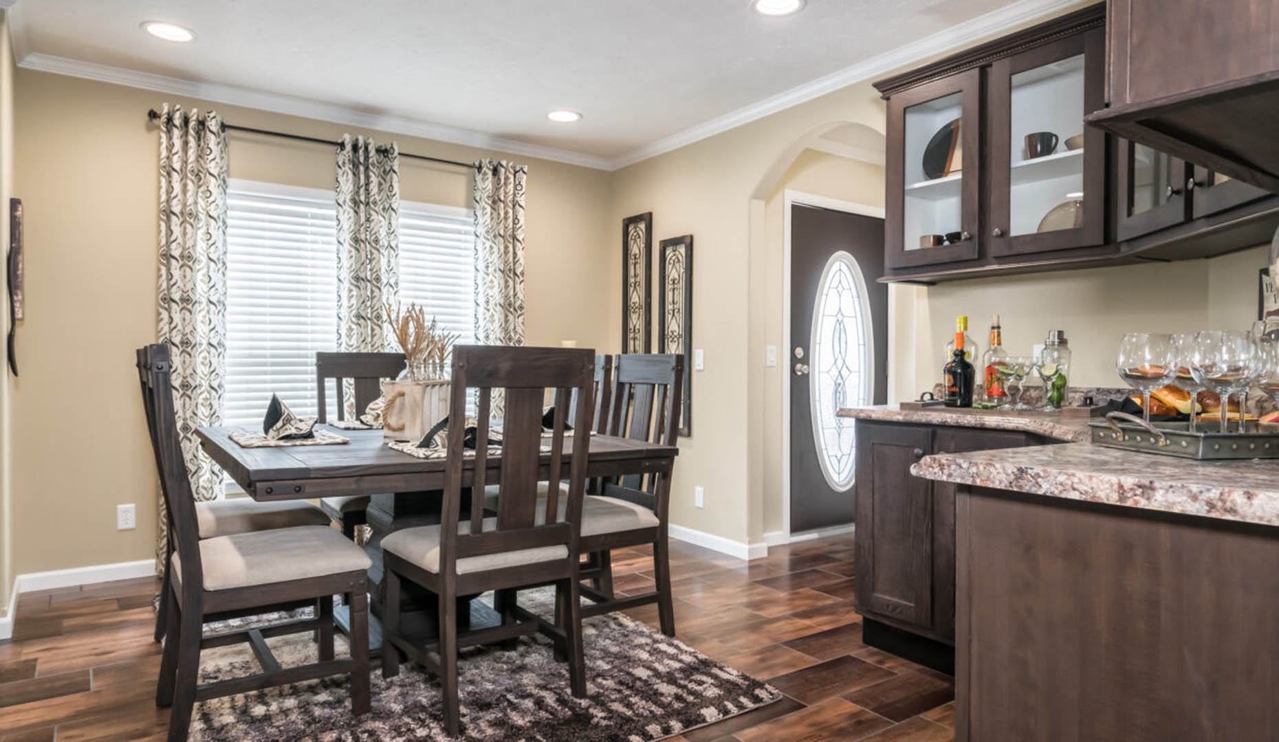 A cozy dining room with a wooden table set for six, surrounded by beige walls. Cabinets display glassware, and patterned curtains hang by the window.