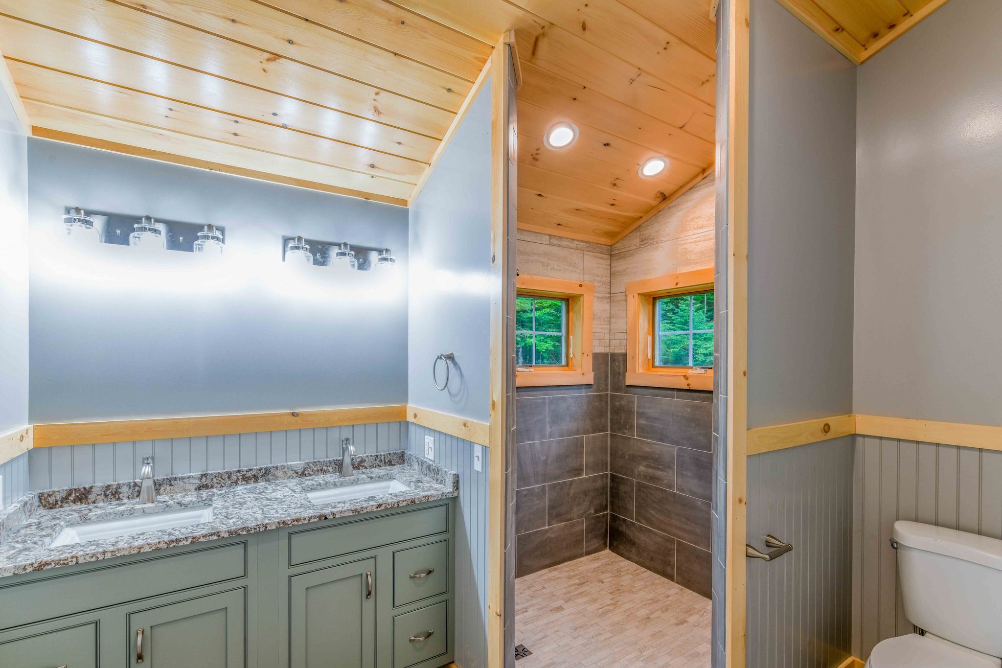 Bright, rustic bathroom with wooden ceilings. Features a dual-sink granite vanity, large tiled shower, and toilet. Natural light through two small windows.