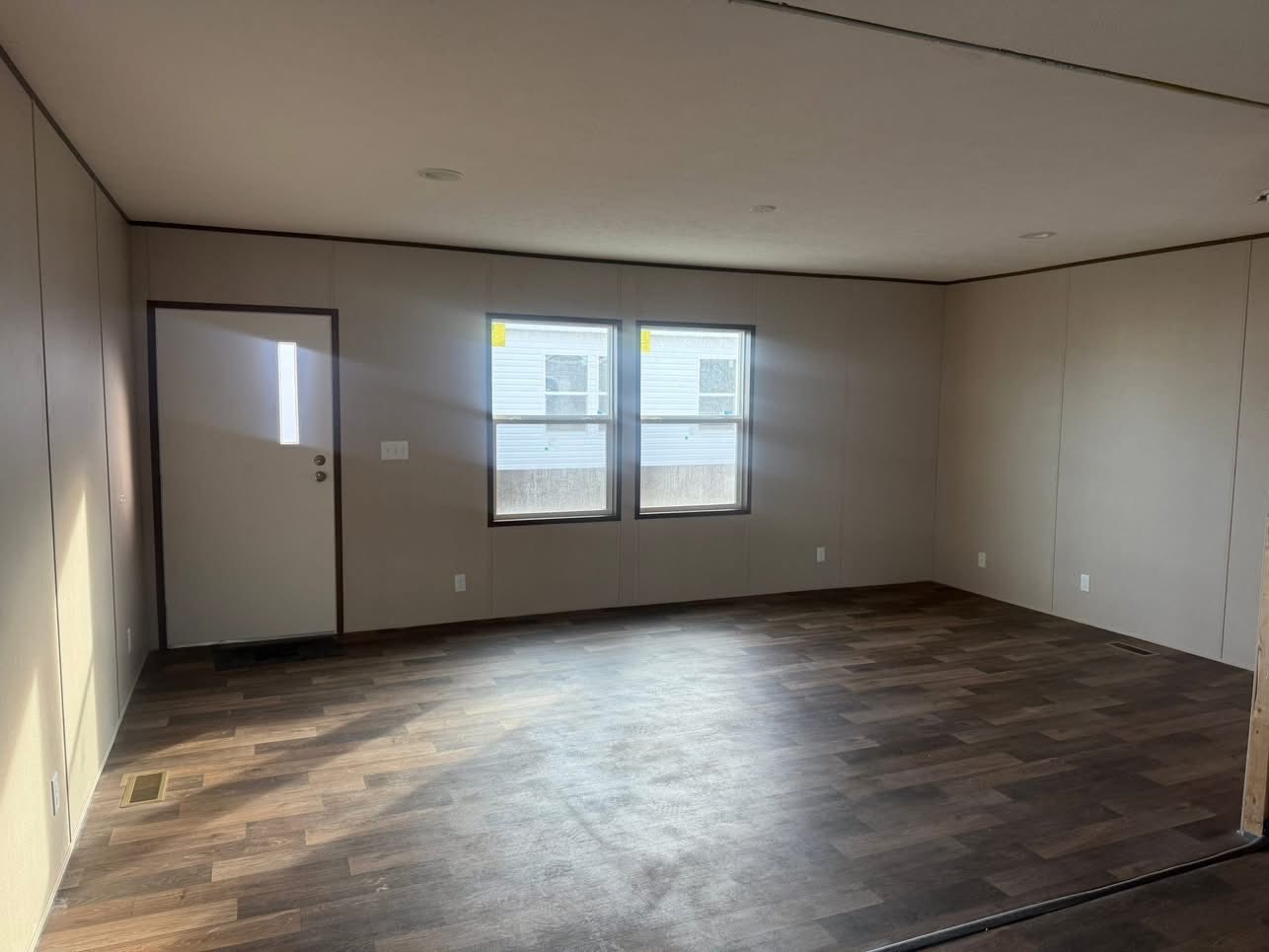 An empty room with beige walls, two large windows, and a door. The wood laminate flooring reflects sunlight, creating a bright and open atmosphere.