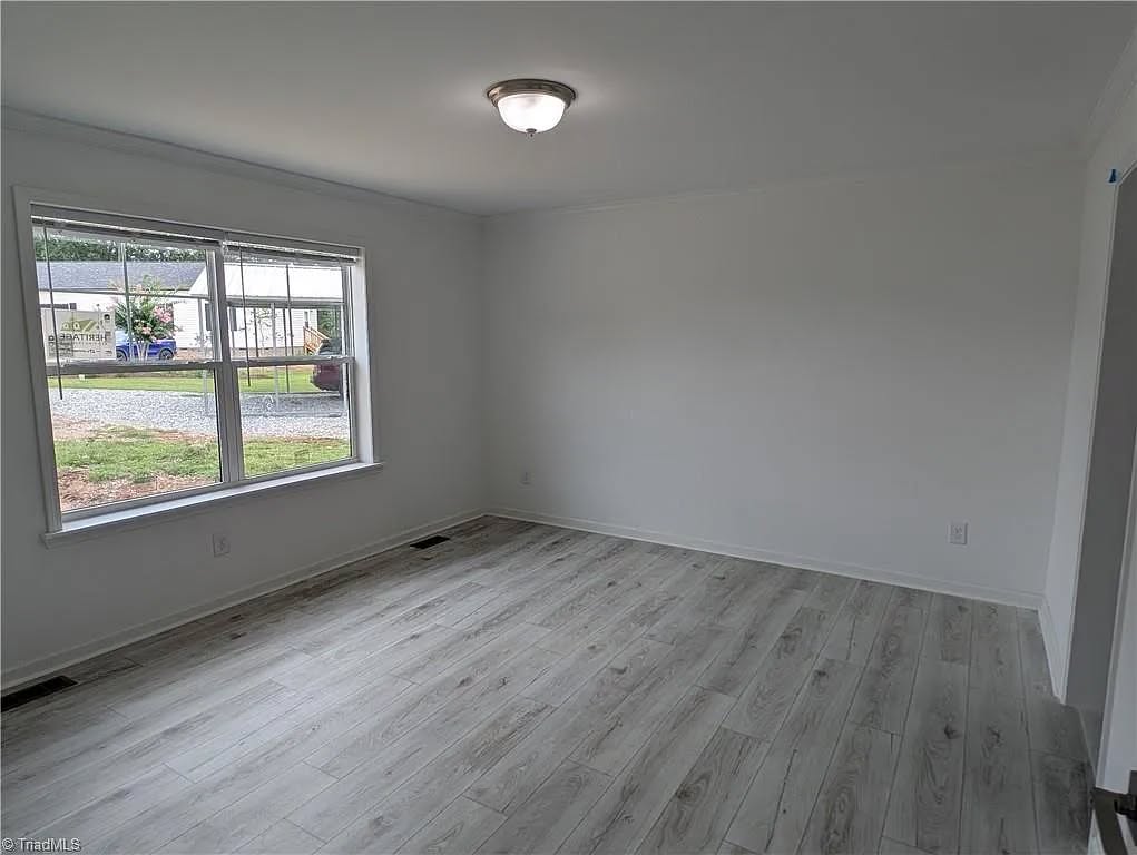 A bright, empty room with light gray wood flooring and white walls. A large window on the left offers a view of a grassy yard and parked cars.