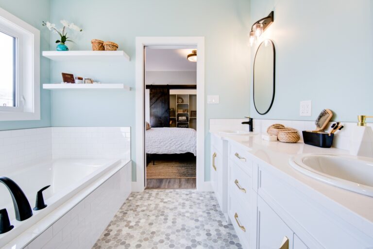 Bright bathroom with pastel blue walls and white fixtures. Features a bathtub, double sinks, round mirror, and open doorway to a cozy bedroom.