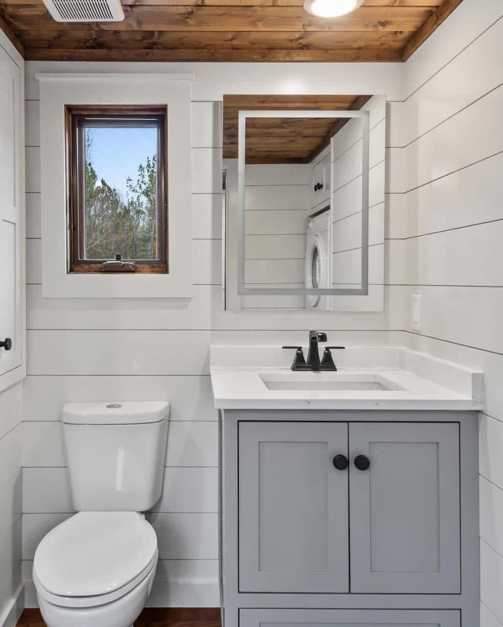 Compact bathroom featuring white shiplap walls, a wooden ceiling, a single small window, a light gray vanity with a sink, and a white toilet. The atmosphere is cozy.