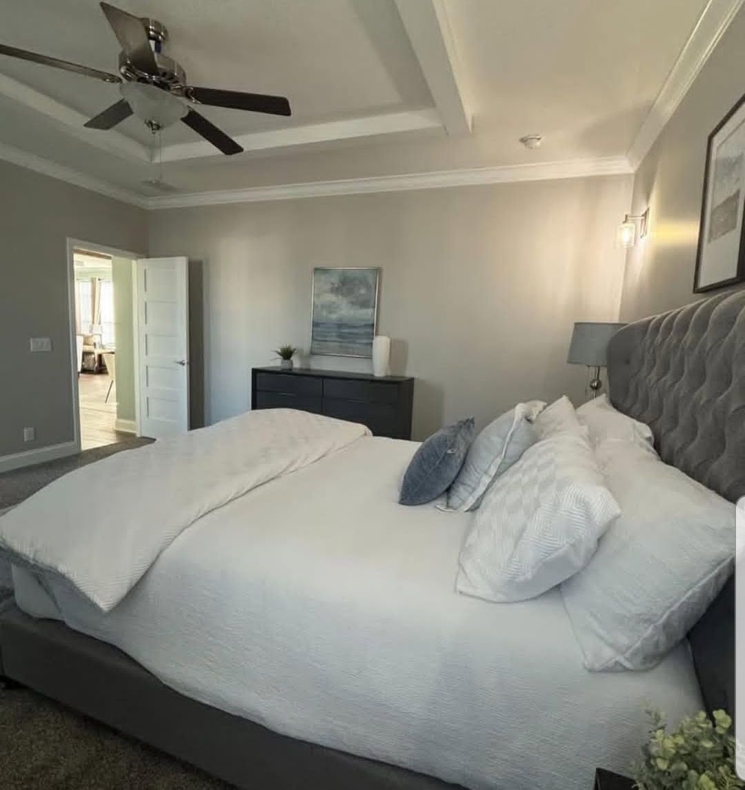 Spacious bedroom with a large, neatly made bed, white bedding, and gray tufted headboard. Minimalist decor with a ceiling fan and soft lighting.