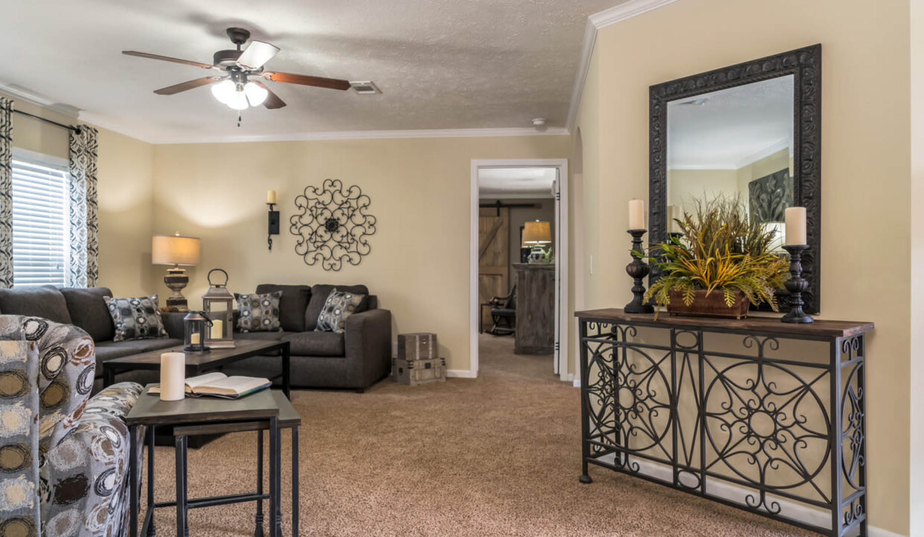 A cozy living room with beige walls, carpeted floor, and brown sectional sofa. Decorative iron elements, large mirror, candles, and plants add warmth.
