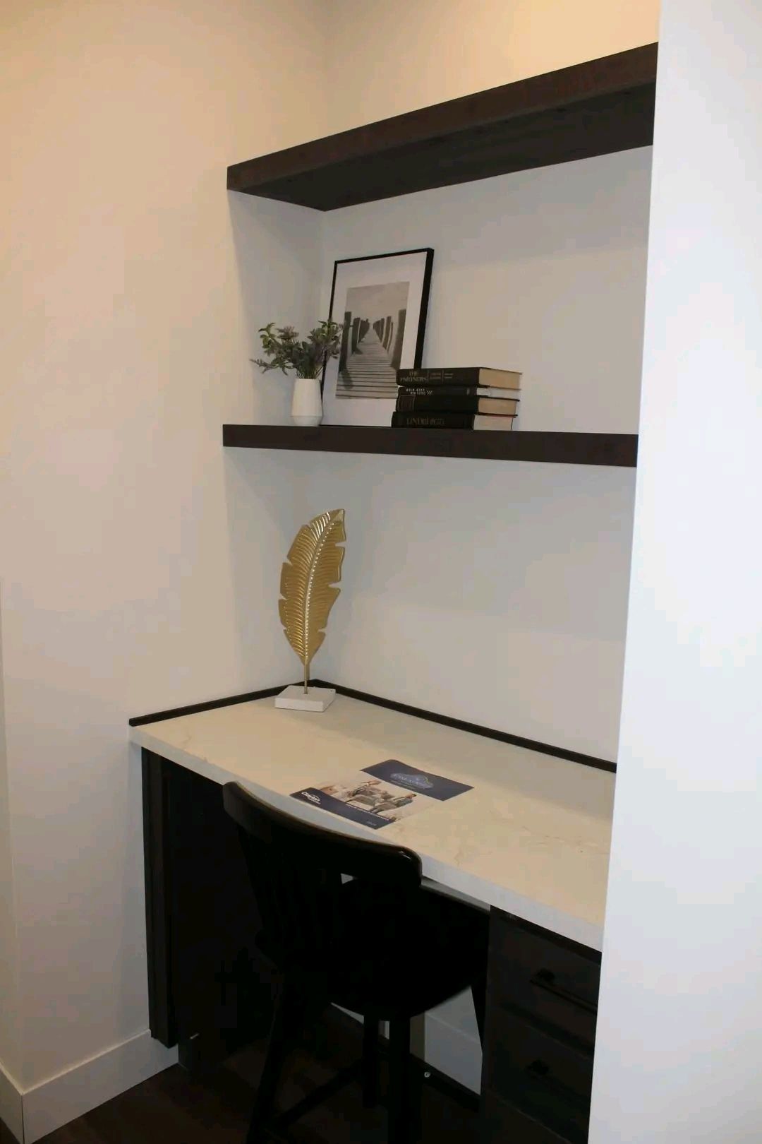 A minimalist home office nook with a white desk, dark chair, and shelving. Decor includes a gold leaf ornament, books, and a framed photo.