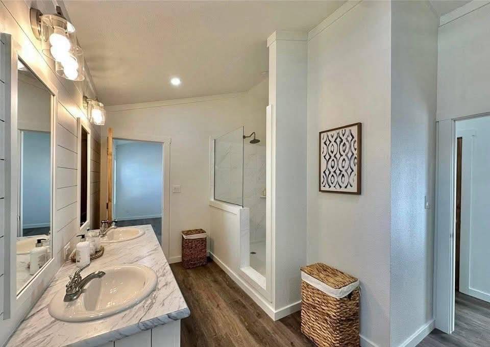 A modern bathroom with double sinks on a marble countertop, a glass shower, and wicker hampers. The space is bright with wooden floors and white walls.