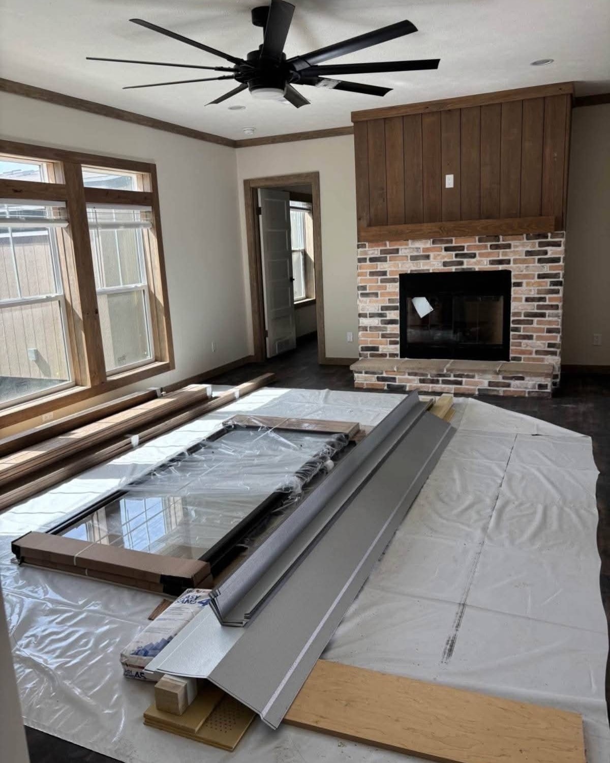 A living room under renovation with building materials on the floor, a ceiling fan above, three tall windows on the left, and a brick fireplace ahead.