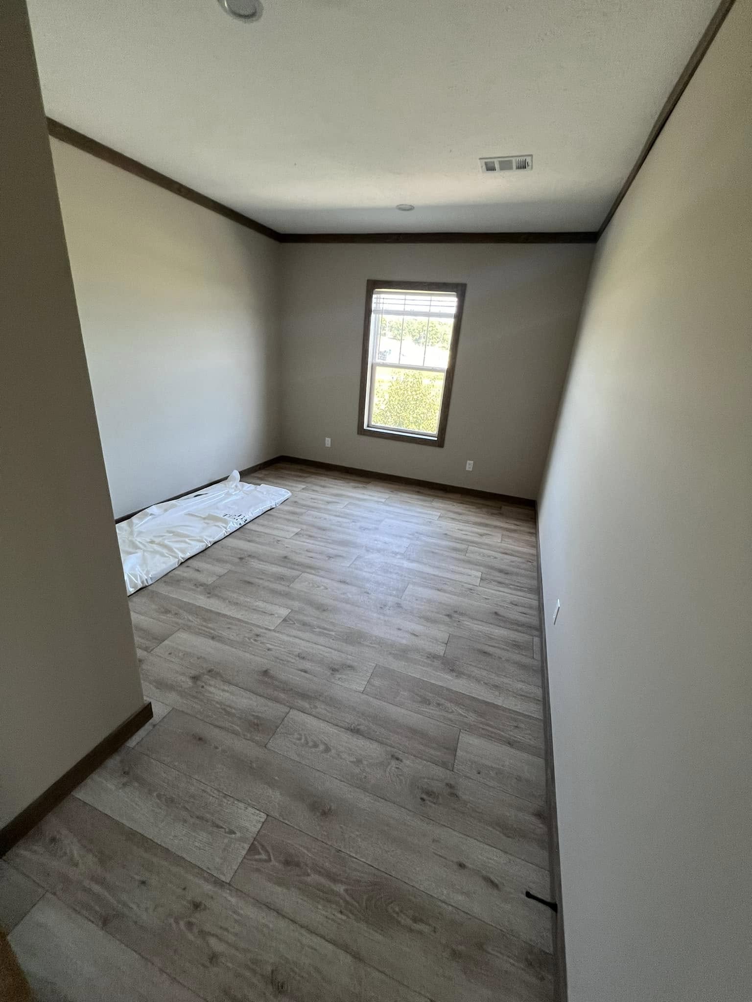Empty room with light wood flooring, beige walls, and a single window letting in daylight. A folded white sheet is placed near the wall. The atmosphere is minimal and calm.