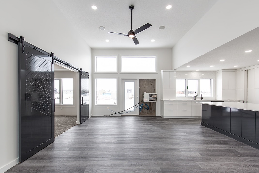Spacious modern interior with dark wood flooring, black sliding barn doors, and large windows. Bright kitchen area on the right, ceiling fan centered.