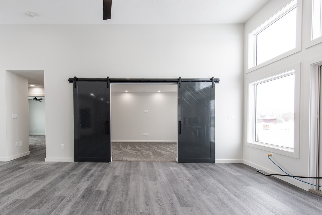 Modern living area with gray hardwood floors and large windows. Black sliding barn doors open to reveal a carpeted room with recessed lighting, creating a sleek, minimalist ambiance.
