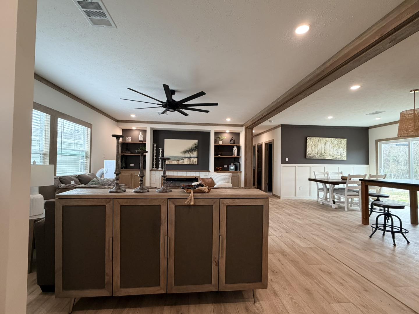 Spacious living area with modern decor, featuring a wood cabinet, ceiling fan, cozy sofa, and large dining table. Soft lighting creates a warm, inviting atmosphere.