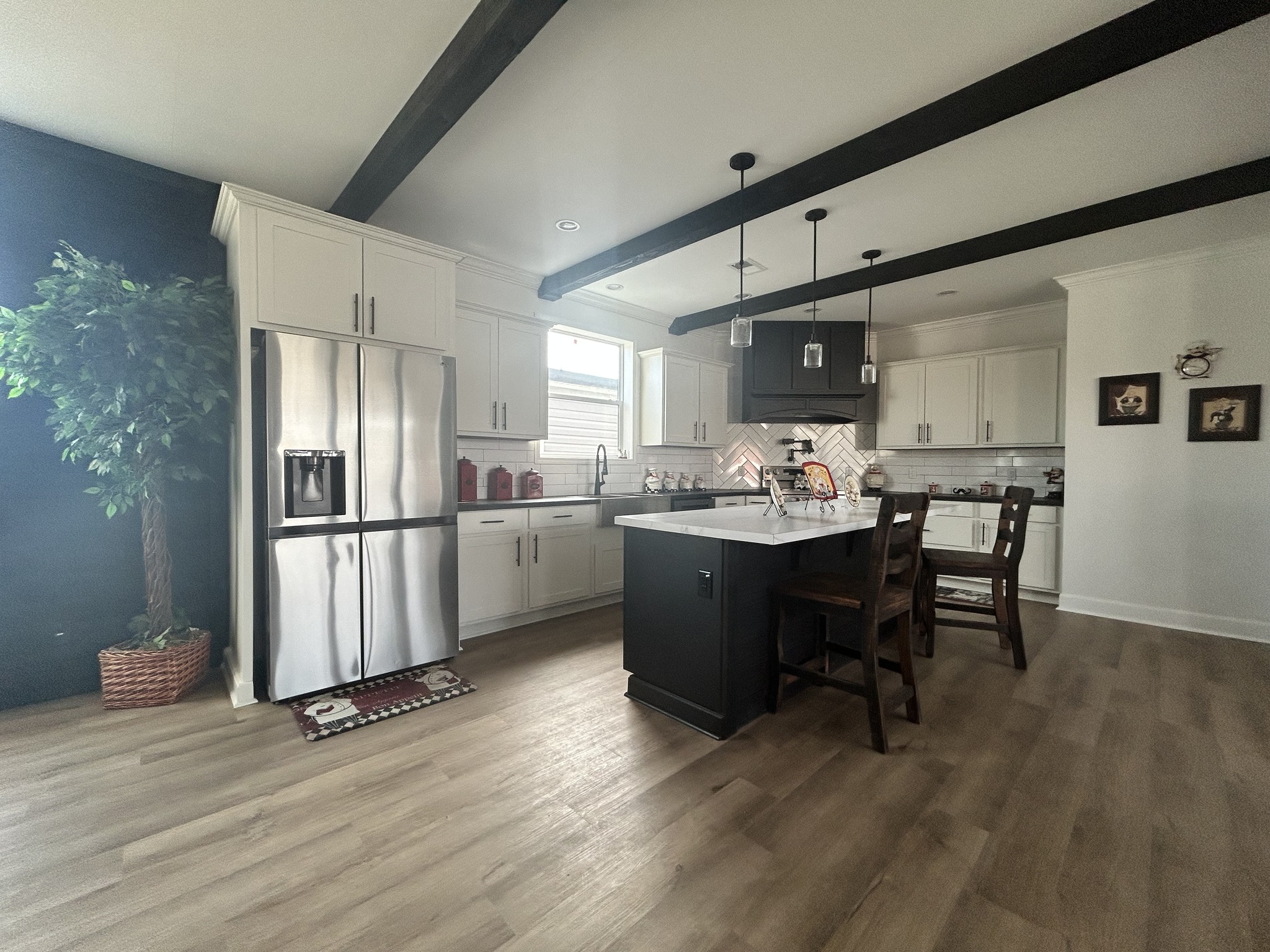 Modern kitchen with white cabinets, stainless steel appliances, and a central island with wooden stools. Black ceiling beams add contrast. A potted plant and two small framed artworks are visible, creating a cozy and elegant ambiance.