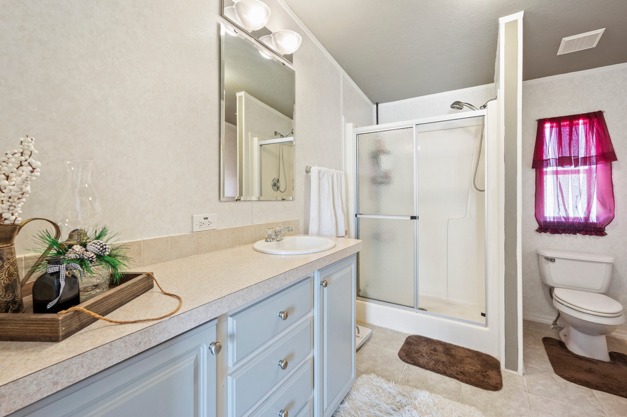 Bright bathroom with a white vanity and mirror, shower with glass doors, and toilet. Decor includes potted plant, brown rugs, and maroon curtains.