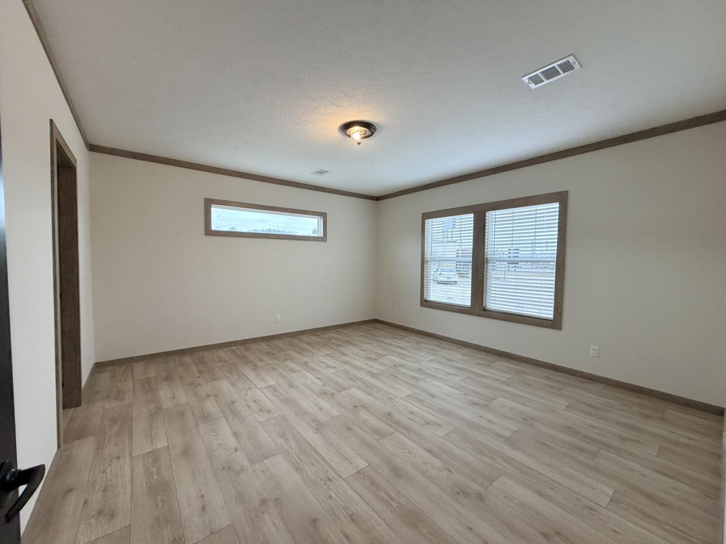 An empty room with light wood flooring and beige walls, featuring a ceiling light. Two windows with blinds allow natural light to enter, creating a calm atmosphere.