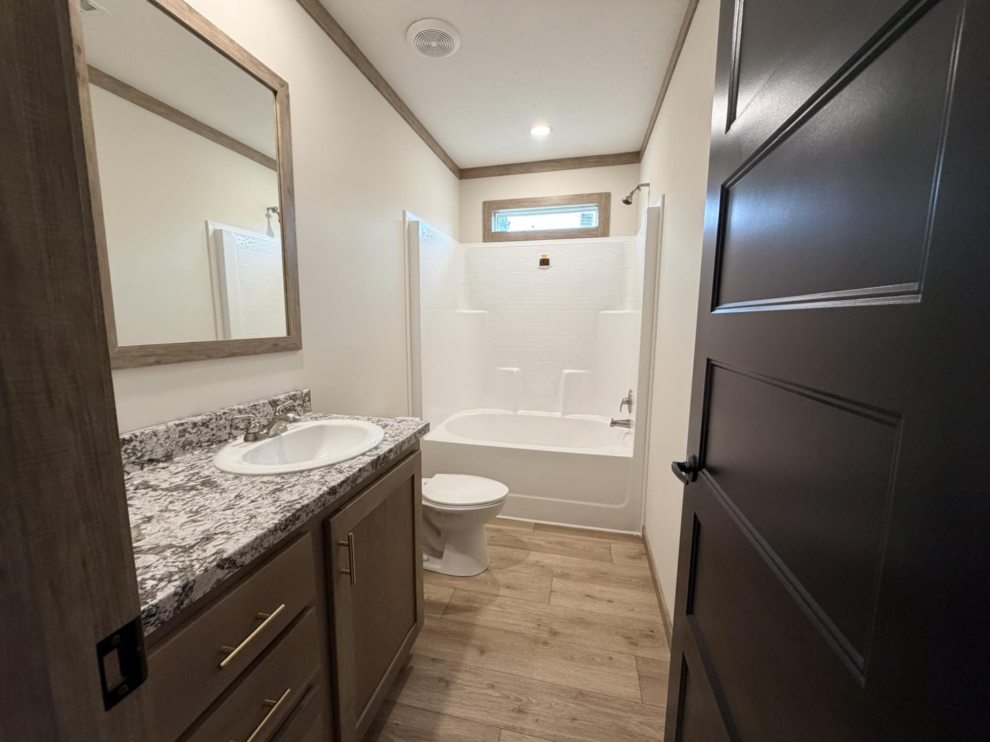 A modern bathroom with light wooden flooring and neutral walls. It features a granite countertop with a sink, a toilet, and a bathtub with a shower.