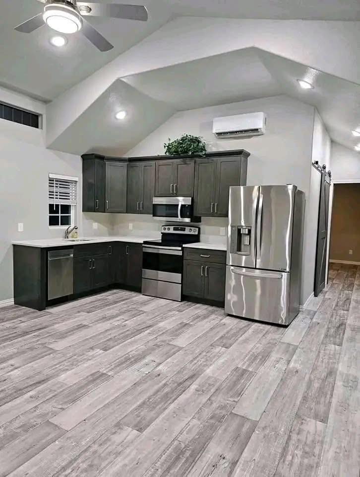 Modern kitchen with dark cabinets, stainless steel appliances including a refrigerator and oven, light wood flooring, and a ceiling fan.