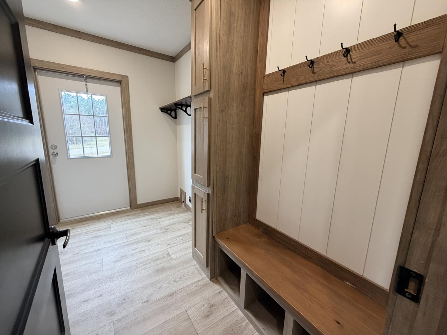 Mudroom with wooden benches and coat hooks along the right wall, light wood flooring, and a door with a window; bright, organized, functional space.