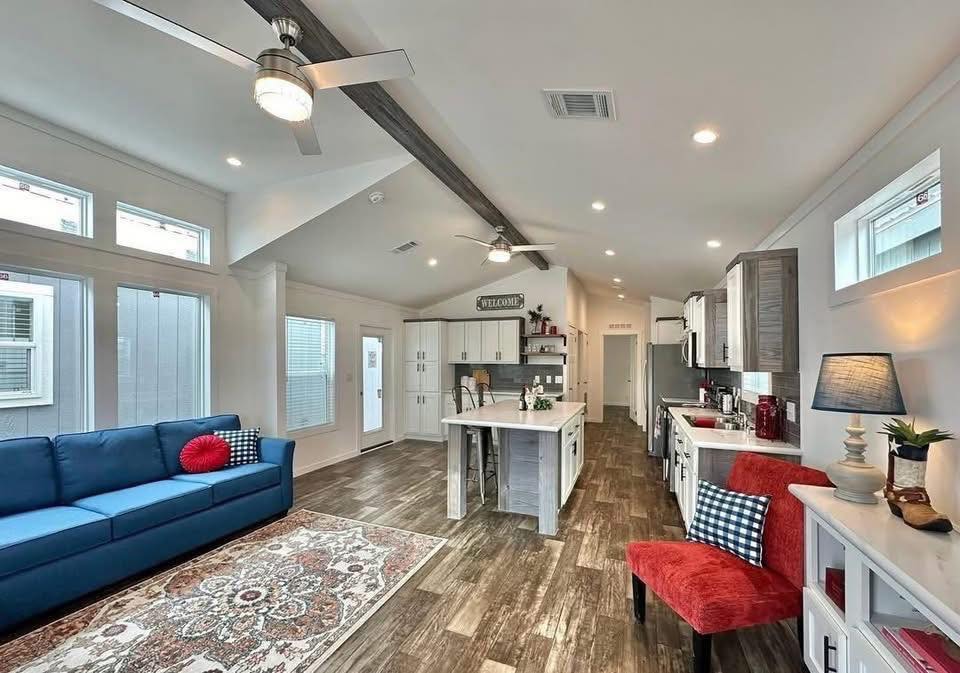 Spacious, modern living area with wooden floors, blue couch, red chair, and kitchen island. Large windows and ceiling fans enhance the airy, welcoming ambiance.