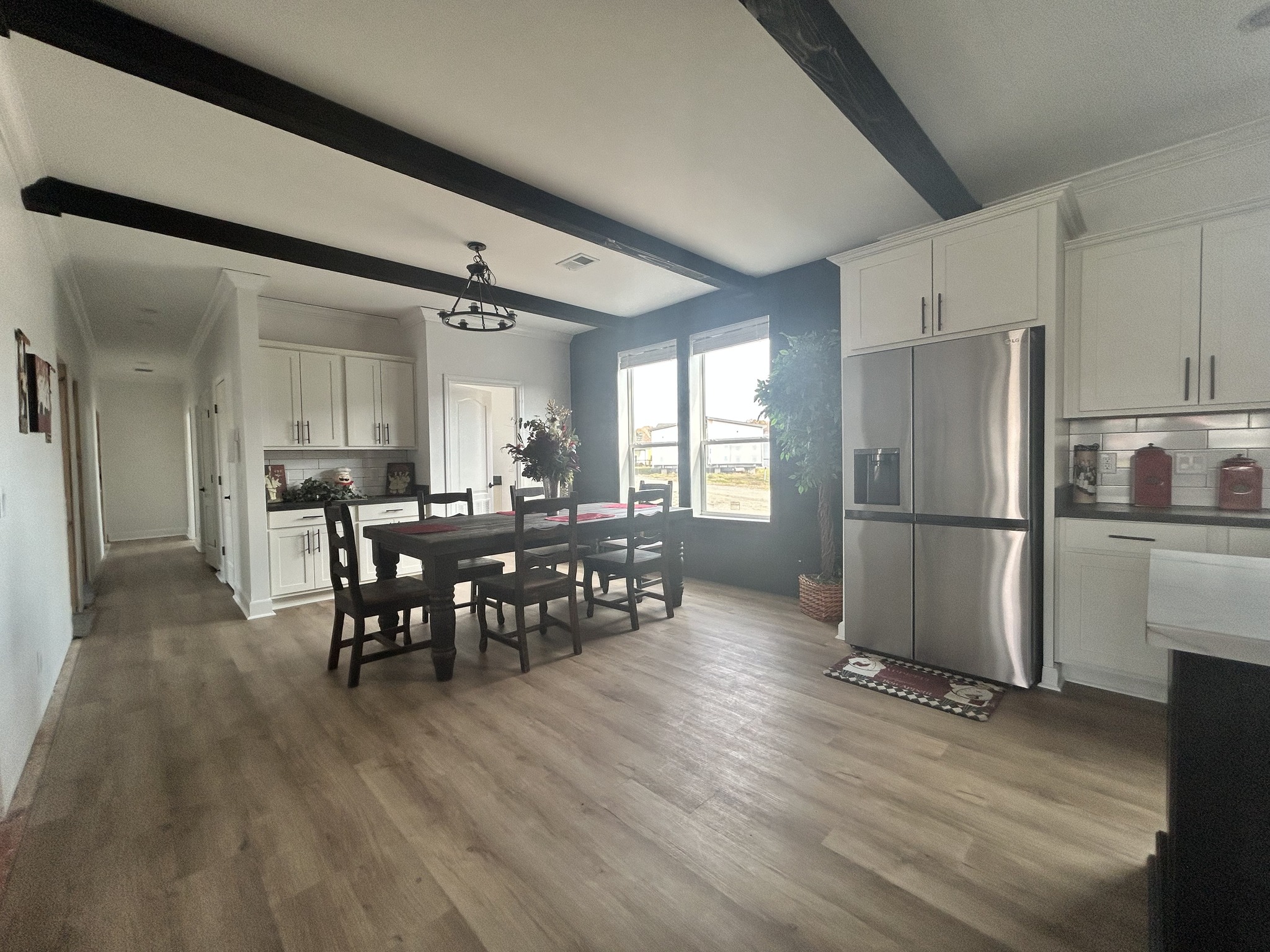 Spacious kitchen-dining area with wooden floors, black beams, and white cabinets. A wooden dining set sits beside large windows. Stainless steel fridge nearby.