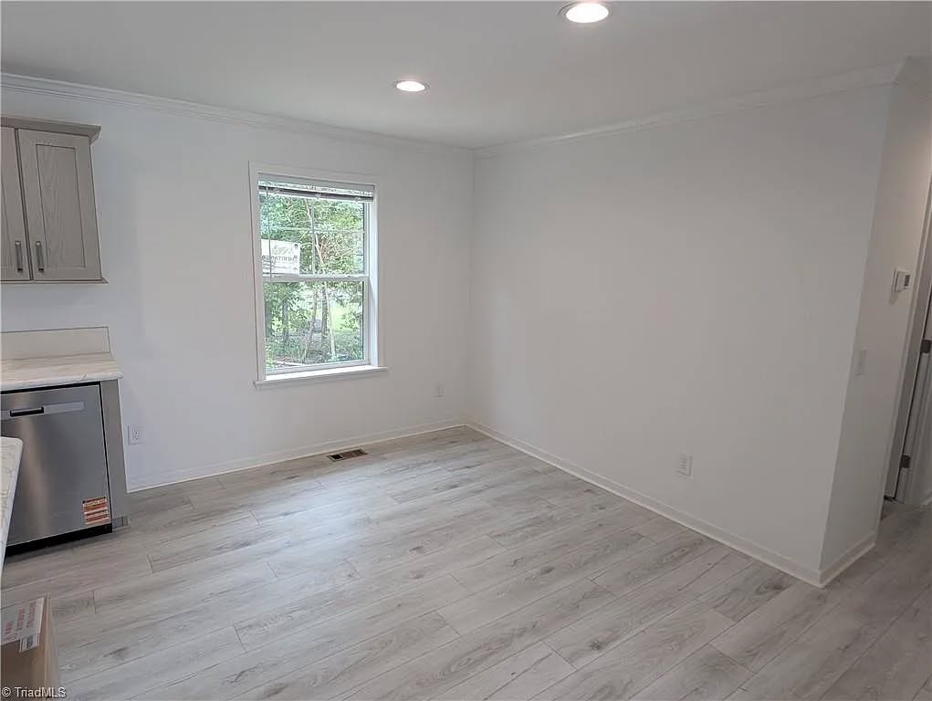 A bright, empty room with light gray wood flooring and white walls. A large window on the left offers a view of a grassy yard and parked cars.