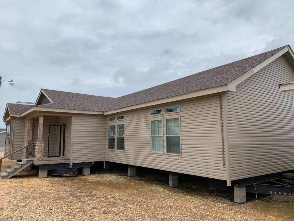 A beige mobile home with a gable roof sits elevated on concrete blocks. It features a small porch, stone steps, and large windows under a cloudy sky.