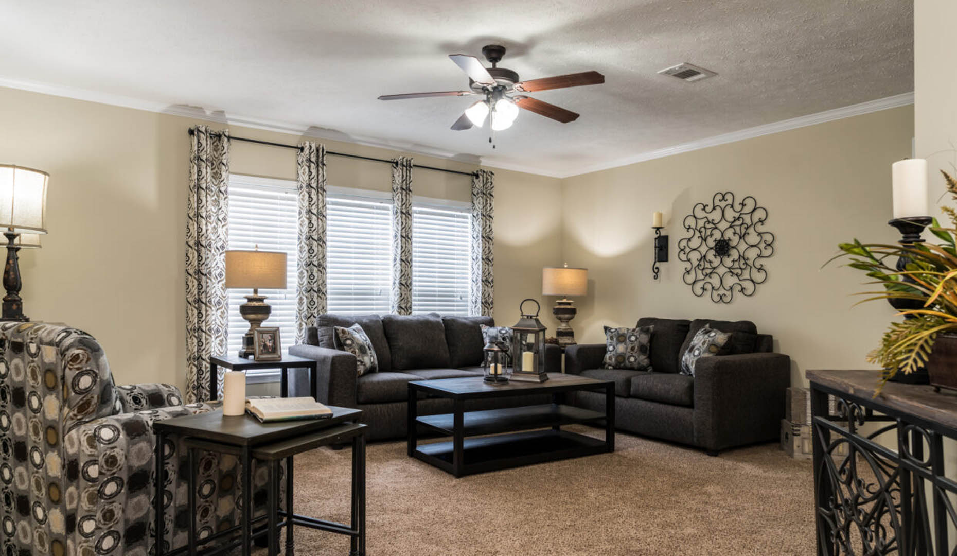 Living room with dark sofas, patterned armchair, coffee table, and lamps. Neutral walls, large windows with drapes, and a ceiling fan create a cozy ambiance.