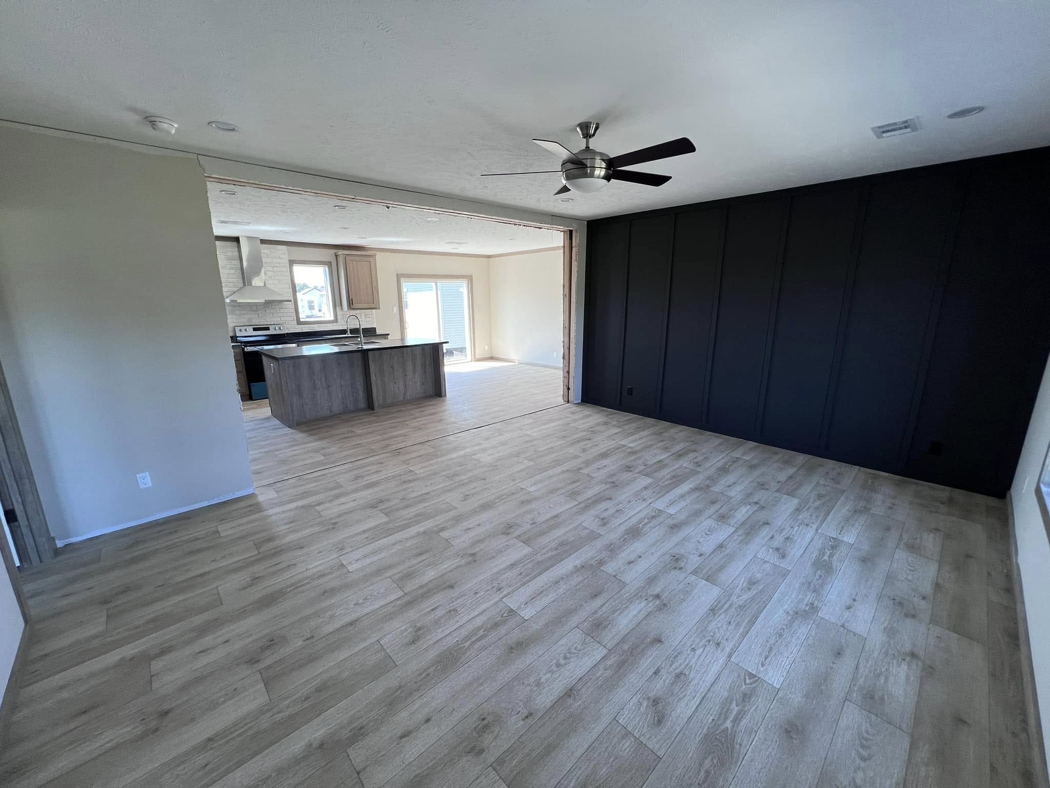 Spacious room with light wood flooring extending to an open kitchen with an island. A dark accent wall contrasts with a ceiling fan above, creating a modern feel.