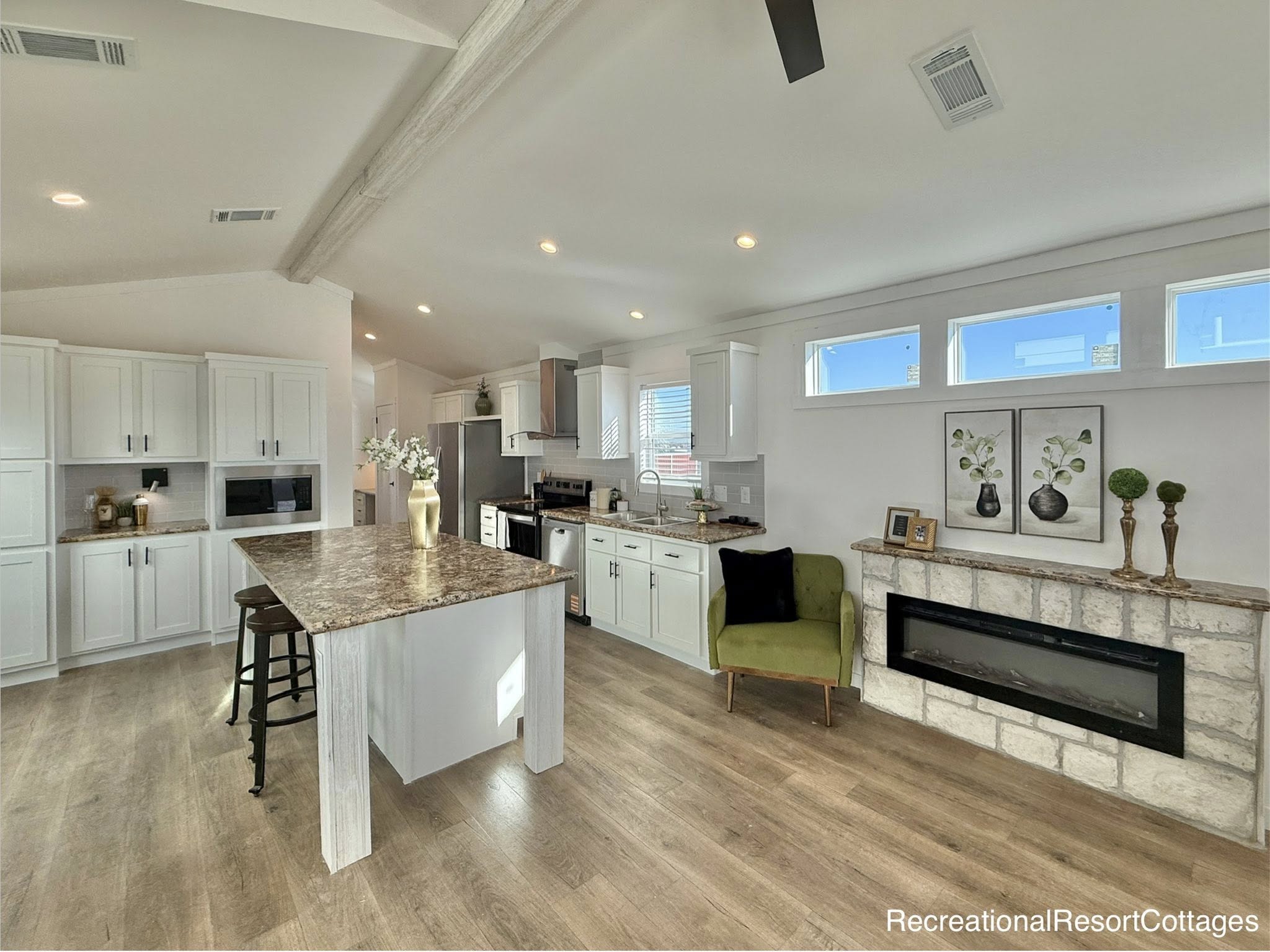 Bright, modern kitchen with white cabinets, granite island, and two stools. Features a cozy green chair, wall art, and a stone fireplace with a sleek design.