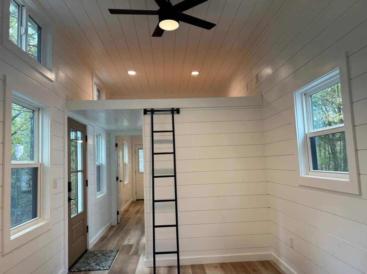 Bright tiny house interior with white shiplap walls, high ceiling, and loft accessed by a ladder. Large windows provide natural light. Cozy and modern.