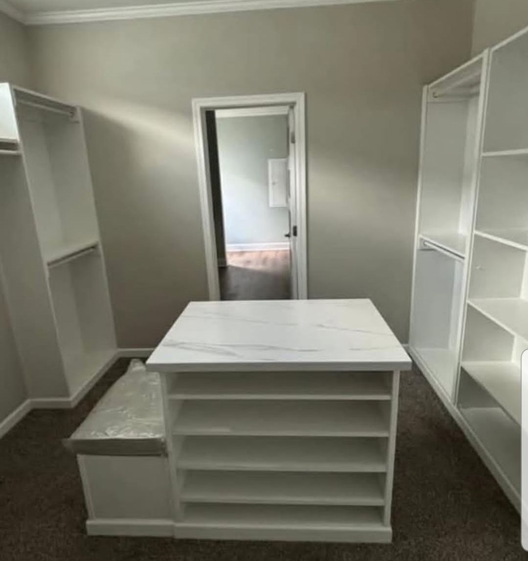 A spacious walk-in closet with white shelves and an island in the center. Minimalistic design, neutral tones, and a tidy, organized feel.